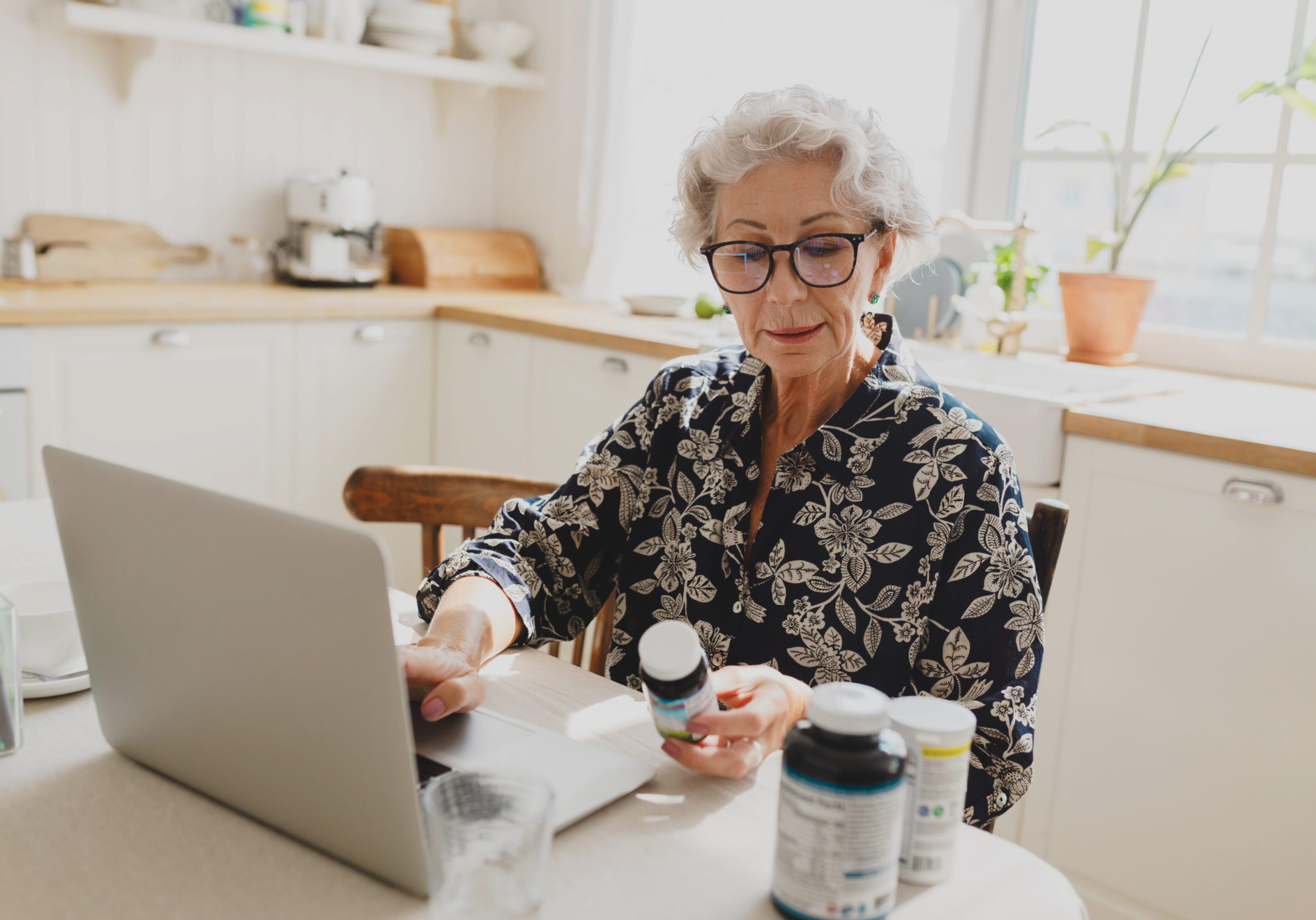 Side view of senior gray-haired female in glasses examining name of food supplement holding bottle, ordering new vitamin on market place or online pharmacy, sitting at kitchen table using laptop A woman sitting at the table with pills and medicine.